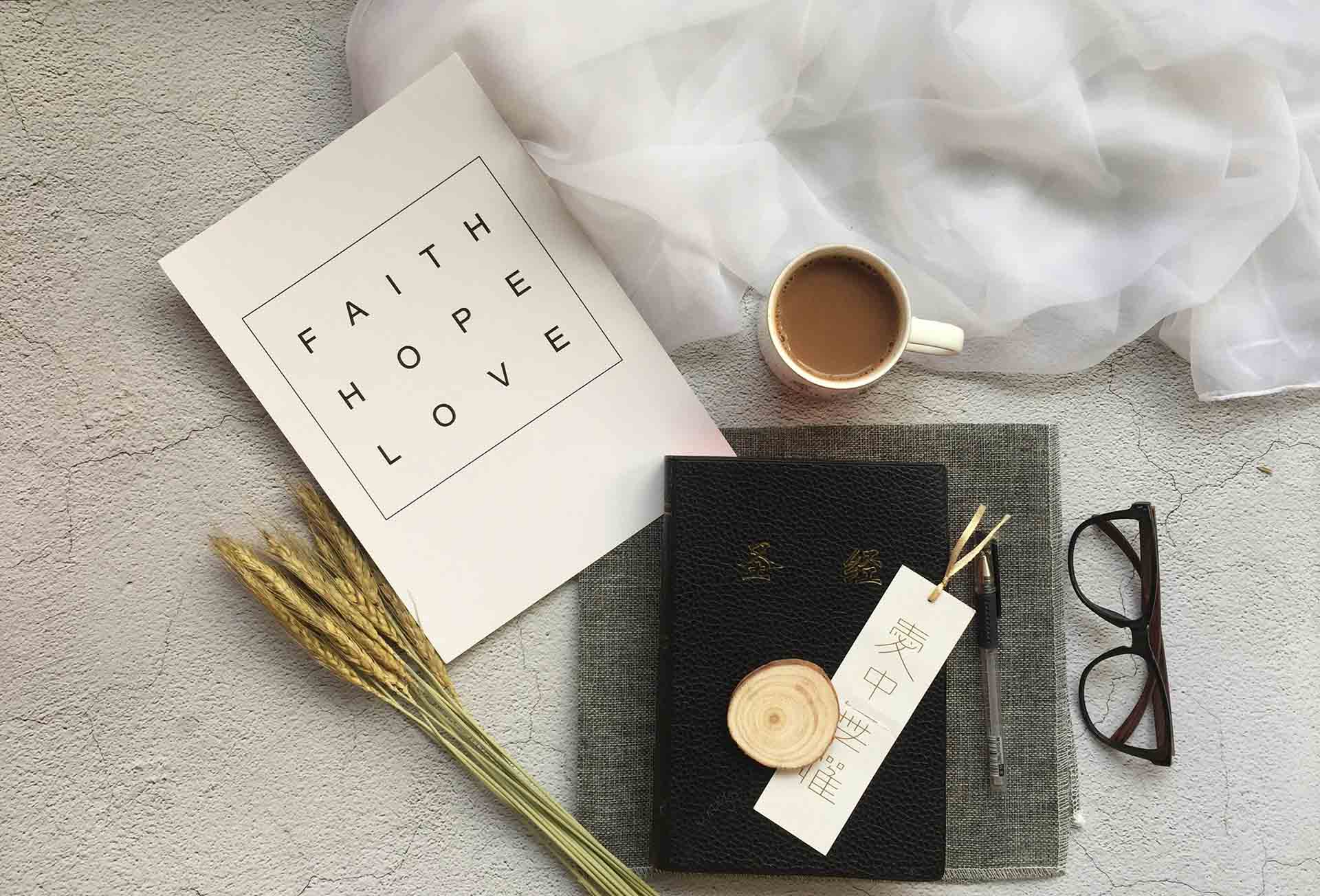 A piece of paper that written of Faith, Hope, Love, a cup of coffee, a glasses, a bookmark on top of a bible